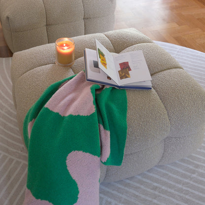 Norah Boucle Ottoman in Warm Beige, a square, tufted piece in boucle fabric. Topped with a book and lit candle, it rests on a patterned rug, offering cozy sophistication.