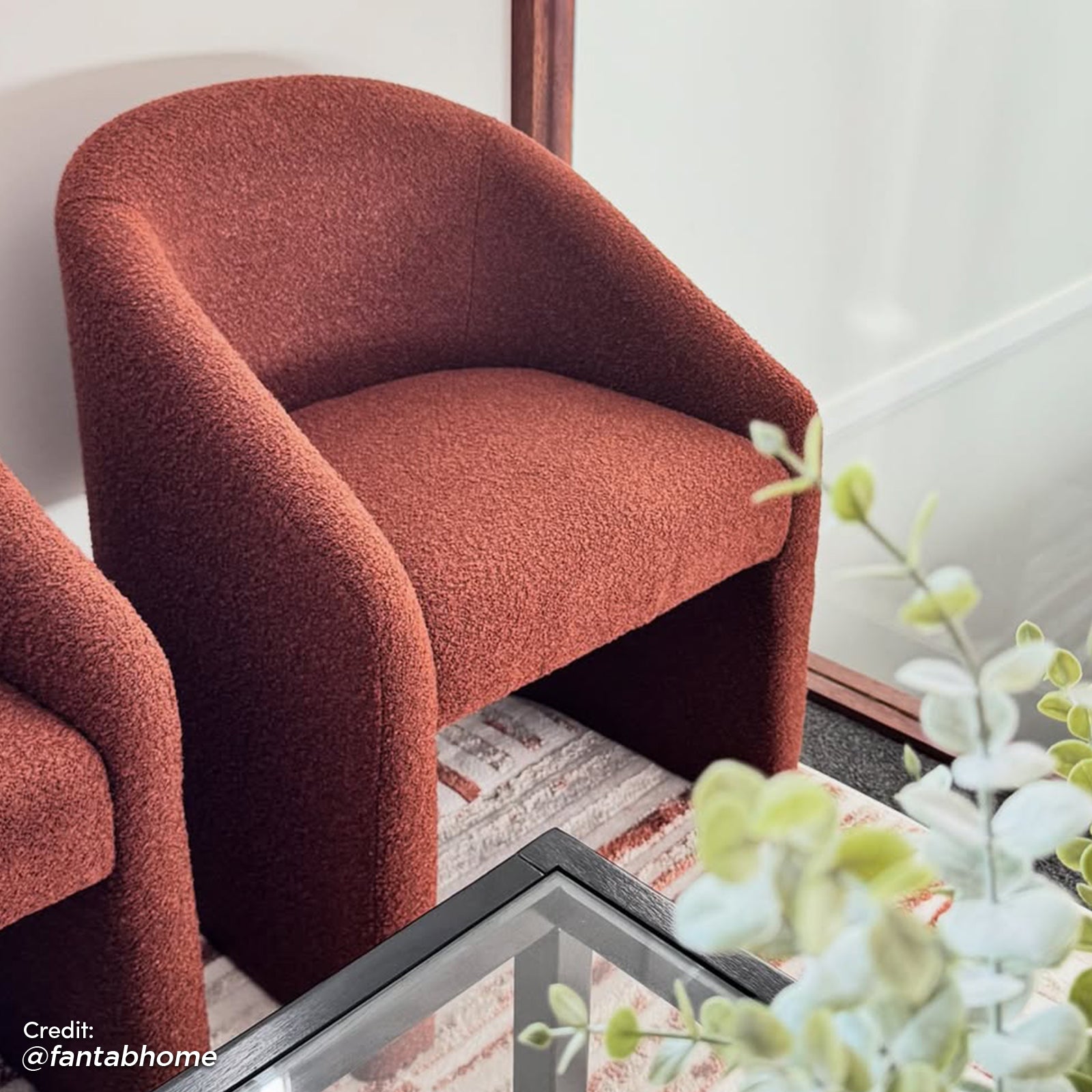 Nuve Bouclé Upholstered Accent Armchair in Rust Brown, featuring a curved silhouette with textured bouclé fabric, positioned on a striped rug beside a black-framed glass coffee table and eucalyptus sprigs.