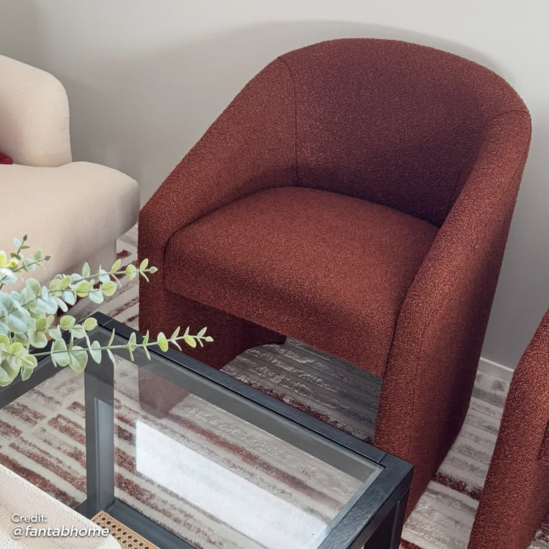 Nuve Bouclé Upholstered Accent Armchair in Rust Brown, featuring a curved silhouette and rich texture, rests on a striped rug beside a cream-colored armchair and a sleek coffee table.