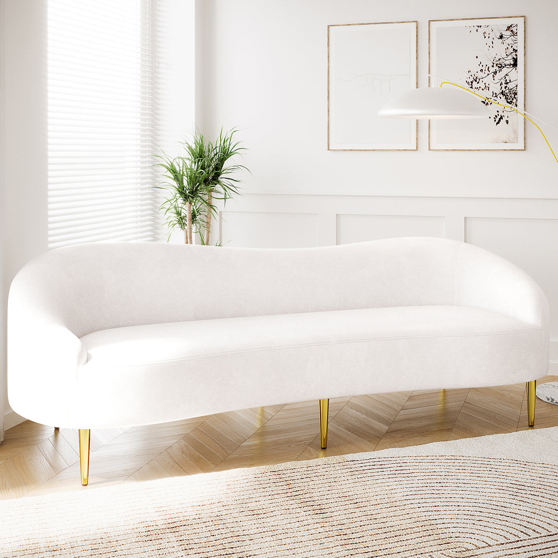Prima 3 Seater Velvet Sofa in Cream with slender brass legs, set in a minimalist living room with wood floors, a neutral rug, and a potted plant beside a window.