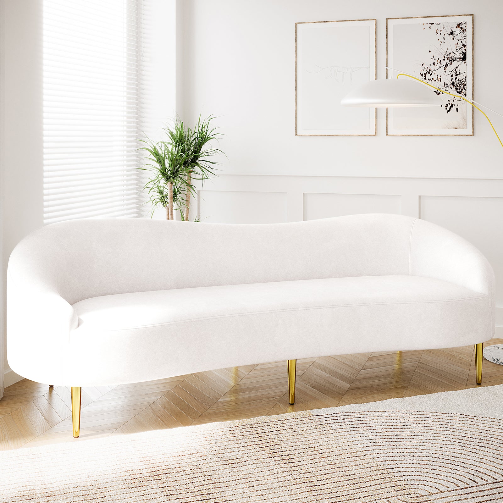 Prima 3 Seater Velvet Sofa in Cream with slender brass legs, set in a minimalist living room with wood floors, a neutral rug, and a potted plant beside a window.