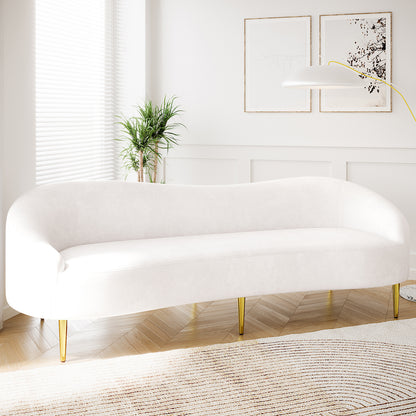 Prima 3 Seater Velvet Sofa in Cream with slender brass legs, set in a minimalist living room with wood floors, a neutral rug, and a potted plant beside a window.