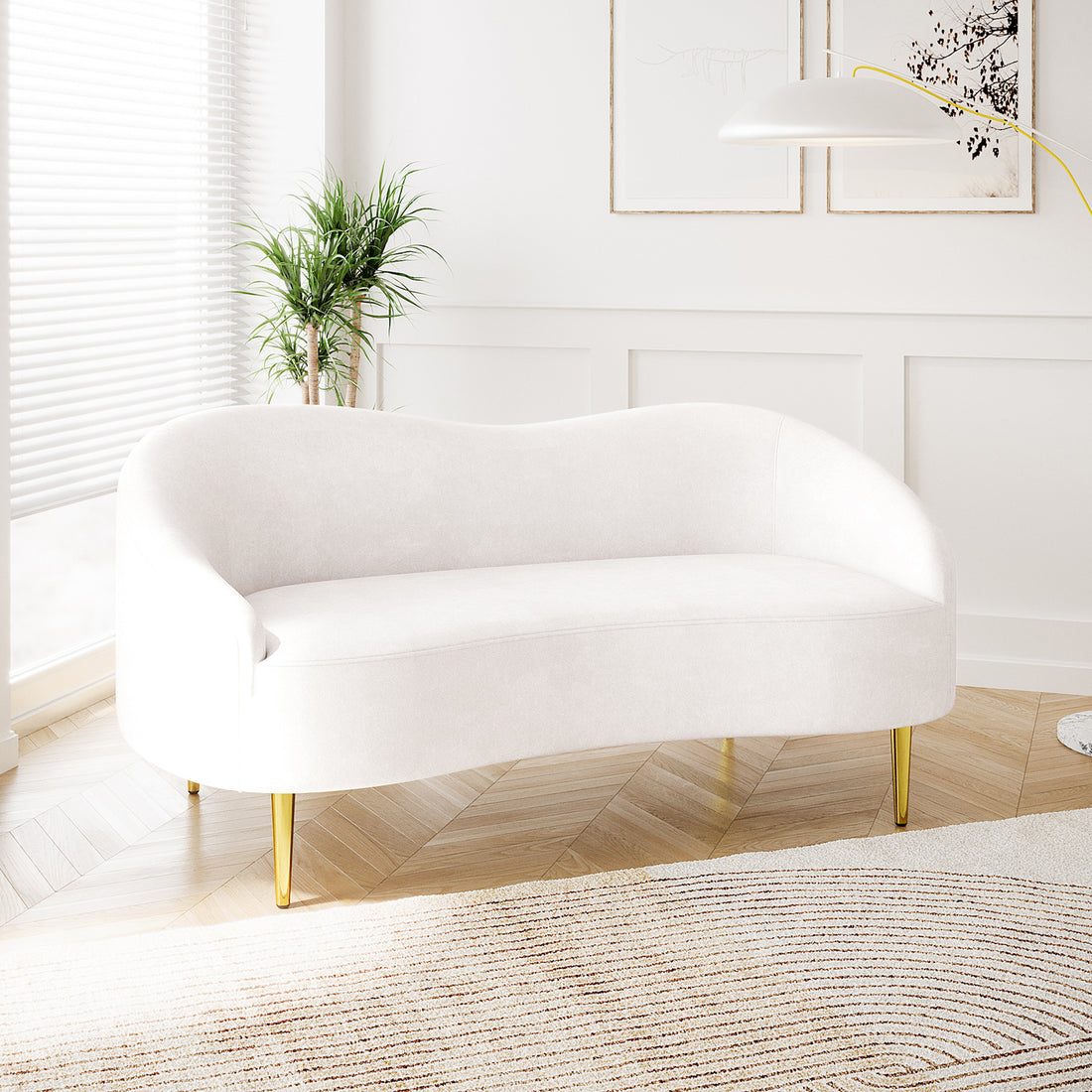 Prima 2 Seater Velvet Sofa in Cream features a sleek, curvy silhouette with gold-toned legs, nestled in a minimalist living room with a herringbone floor, arc lamp, and potted plant.