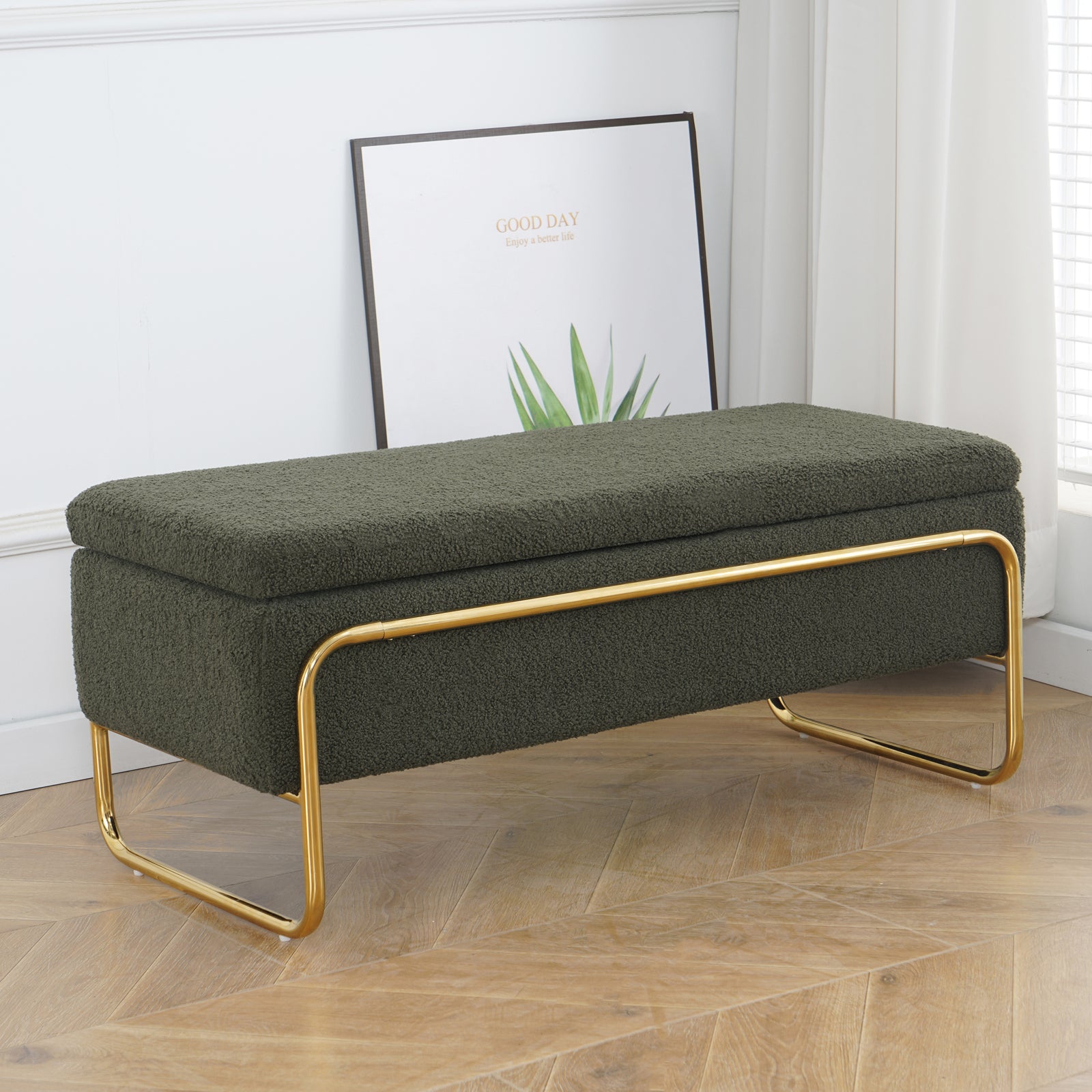 Riane Boucle Storage Ottoman in Green features a rectangular design with boucle upholstery and gold U-shaped legs, set on a wood floor, complemented by a plant-themed print in the background.