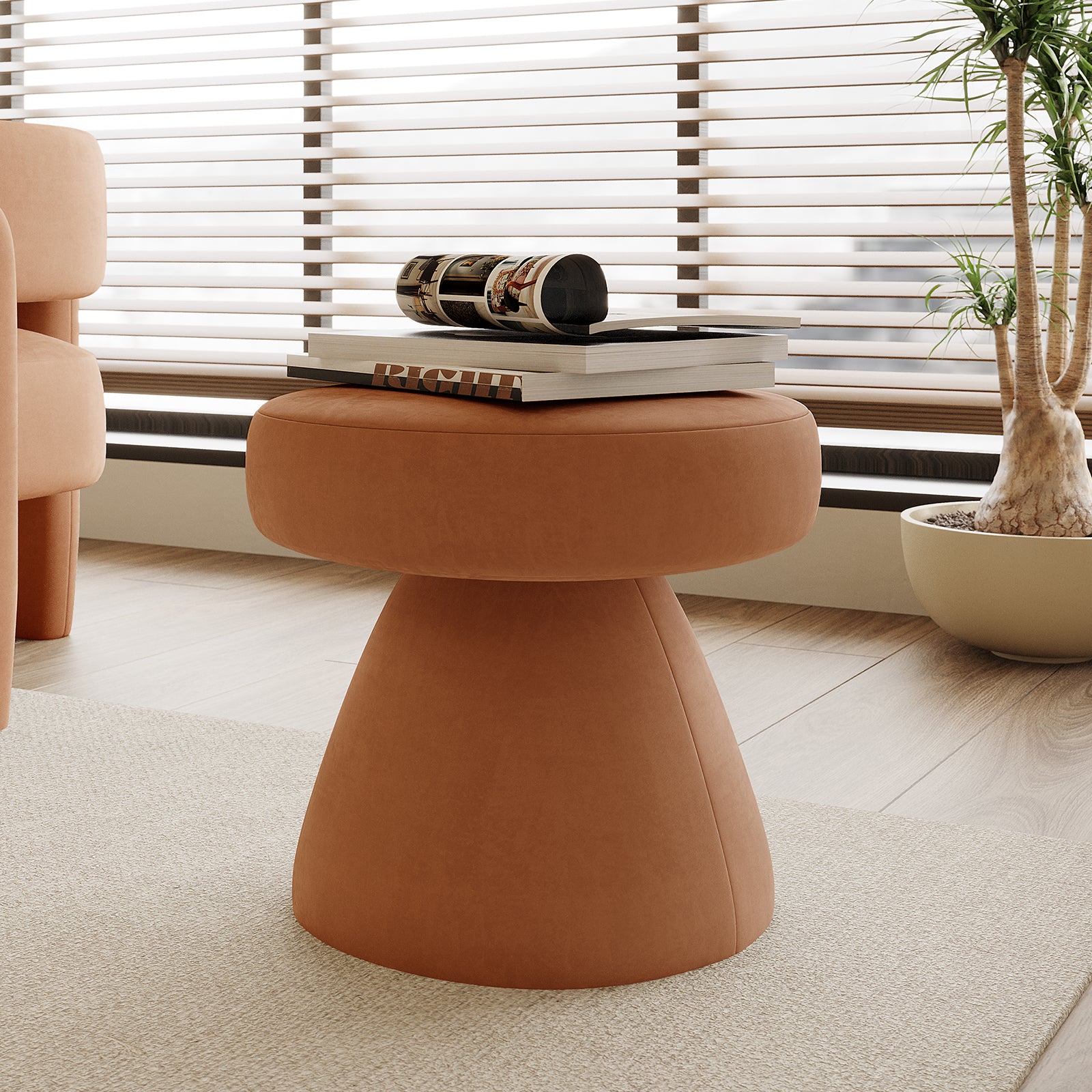 Sadi Velvet Fabric Round Mushroom Ottoman in Burnt Orange, upholstered in terracotta-like material, serves as a stylish centerpiece with art books on top, set against a modern, cozy interior backdrop.