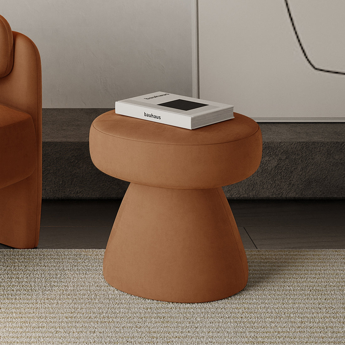Sadi Velvet Fabric Round Mushroom Ottoman in Burnt Orange with rust-brown upholstery, topped with a white book titled “bauhaus,” beside a matching sofa and neutral-tone rug in a modern living space.