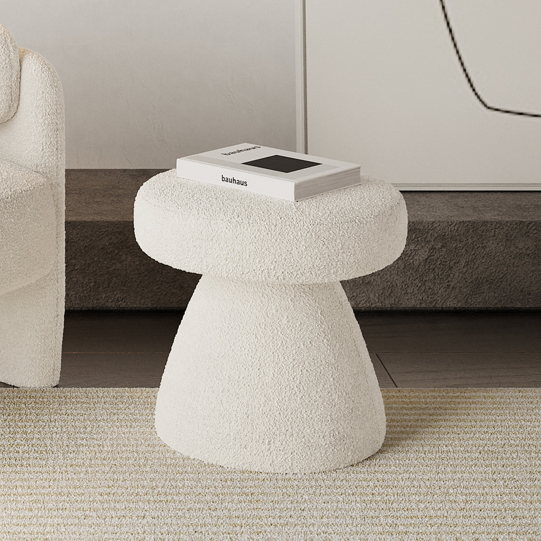 Sadi Bouclé Fabric Round Mushroom Ottoman - Eggshell White, upholstered in off-white boucle, with a bauhaus book on top, situated on a striped rug beside a similarly textured lounge chair.
