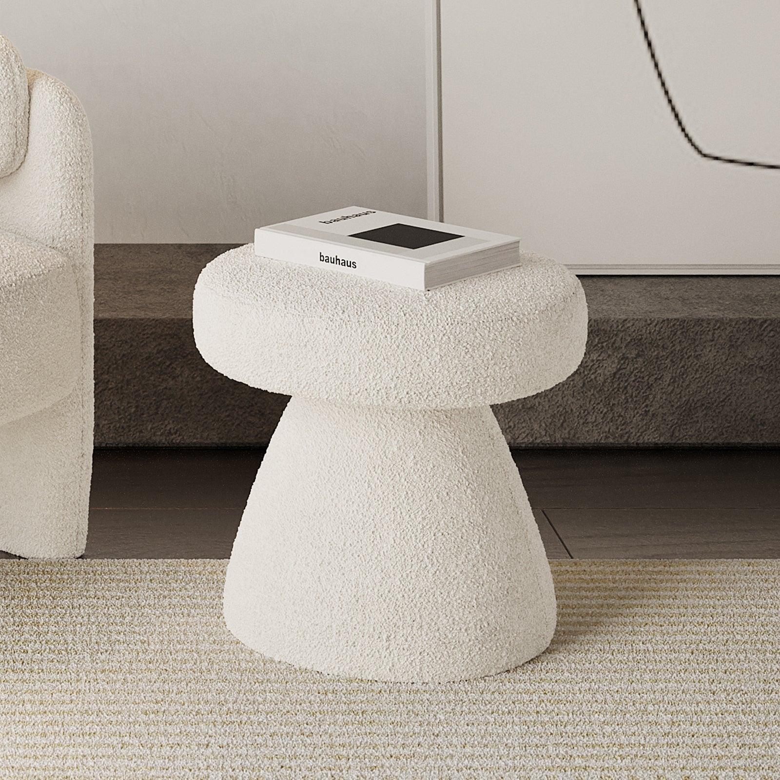 Sadi Bouclé Fabric Round Mushroom Ottoman - Eggshell White, upholstered in off-white boucle, with a bauhaus book on top, situated on a striped rug beside a similarly textured lounge chair.