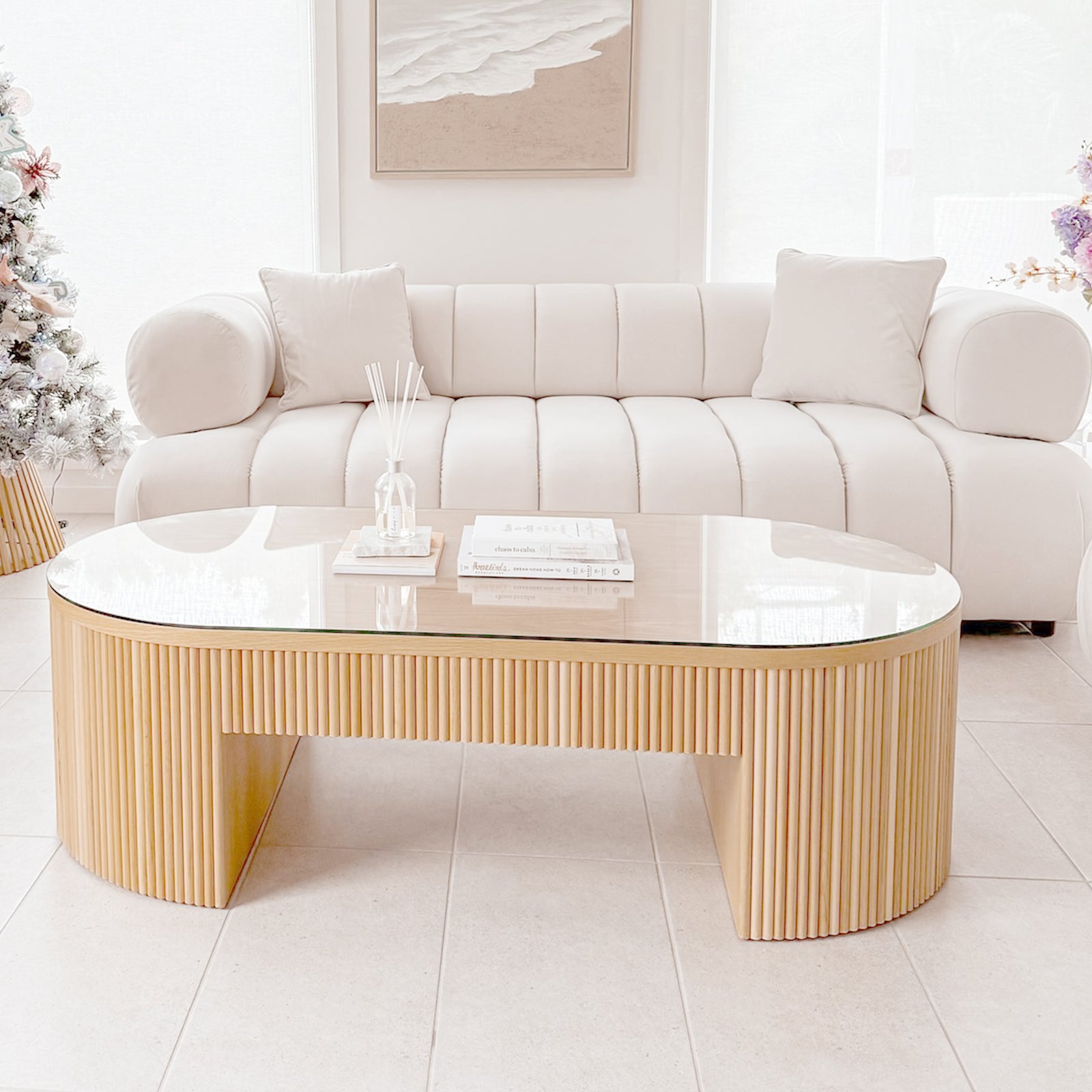 The Samira 3 Seater Velvet Sofa in Cream features plush channel tufting, cylindrical bolster arms, and matching throw pillows, set in a minimalist living room with a light wood slat coffee table.