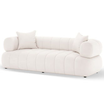 Samira 3 Seater Velvet Sofa in Cream, featuring a low-profile design with off-white velvet upholstery, rounded channel-stitched cushions, cylindrical bolster arms, and matching throw pillows, sits on small, discreet feet.