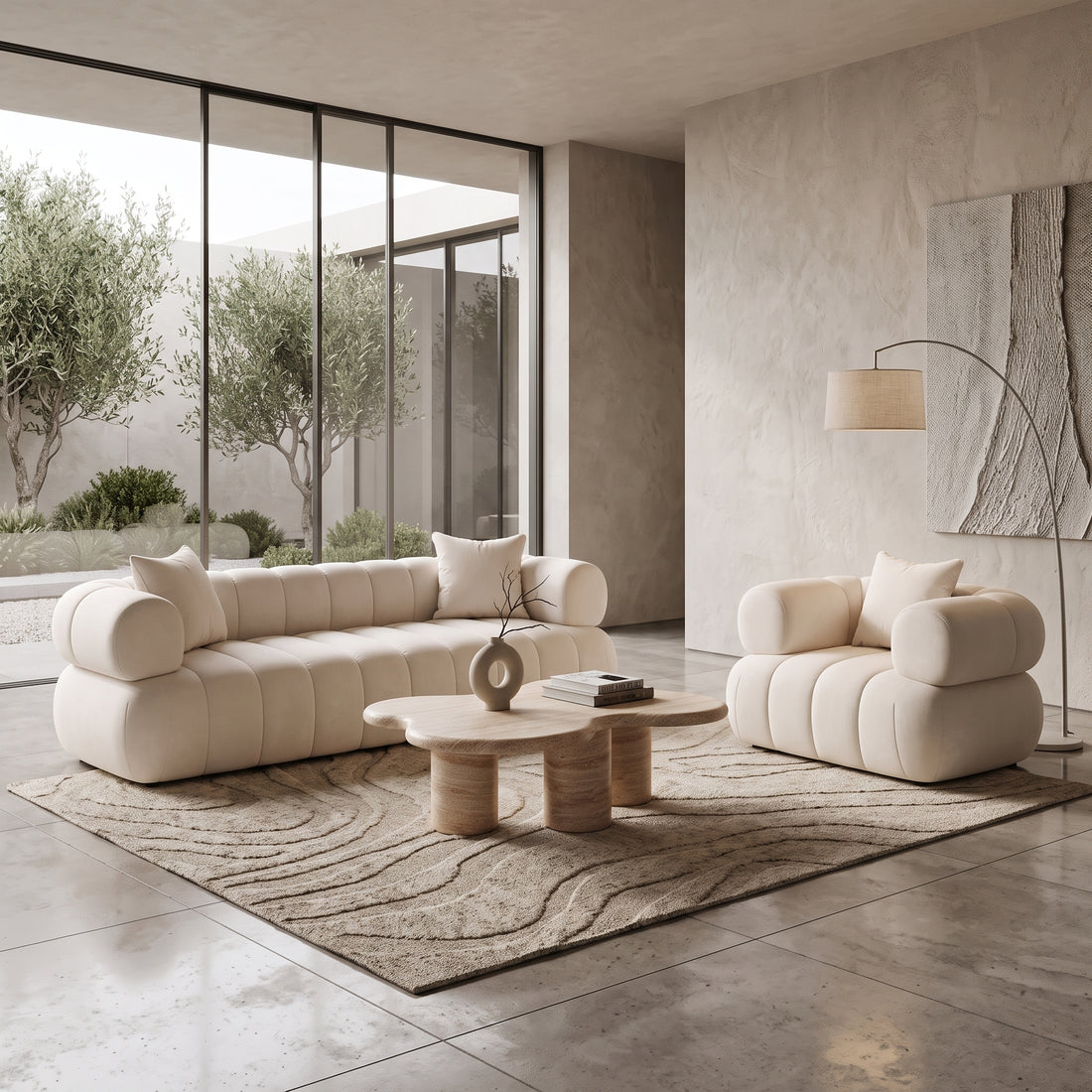A modern living room with a beige channel-tufted sofa and matching armchair, an organic-shaped travertine coffee table on a textured rug, an arched floor lamp, and large windows looking out onto olive trees.