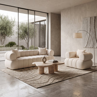 A modern living room with a beige channel-tufted sofa and matching armchair, an organic-shaped travertine coffee table on a textured rug, an arched floor lamp, and large windows looking out onto olive trees.