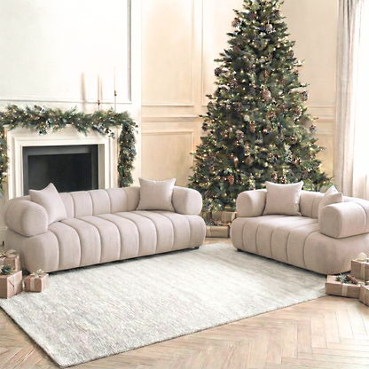 The Samira 5 Seater Velvet Sofa Set in Warm Beige features pale beige scalloped-edge sofas with bolster arms, integrating harmoniously into a softly lit, holiday-decorated living room with a Scandinavian-inspired ambiance.