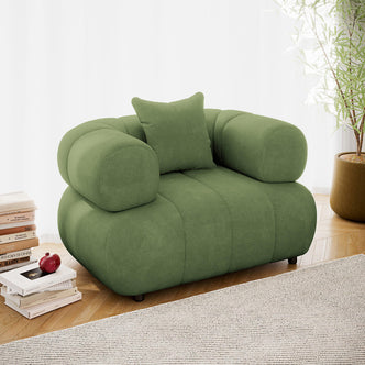 Samira Single Seater Velvet Sofa in Prasinous Green, featuring soft green upholstery, thick armrests, and a matching pillow, set on a herringbone floor with books and a potted plant nearby.