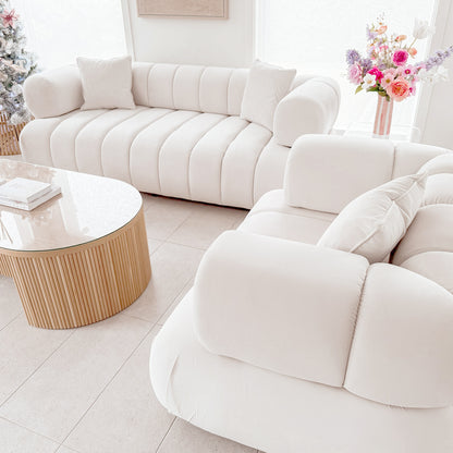 Samira 6 Seater Velvet Sofa Set in Cream features channel-tufted upholstery facing each other, with matching pillows and a round glass-top coffee table between them, creating an inviting, elegant living space.