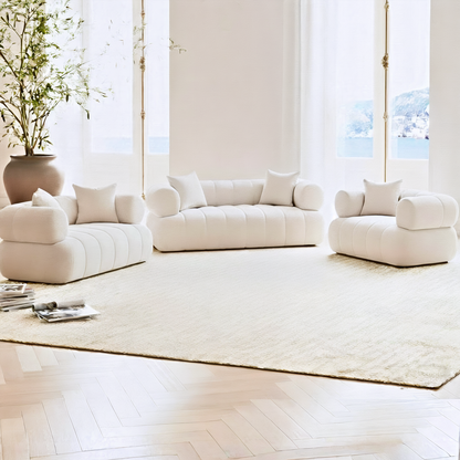 Samira 6 Seater Velvet Sofa Set in Cream, featuring a plush three-piece arrangement, sits in a bright living room with an off-white rug, sheer curtains, and a tall potted plant.