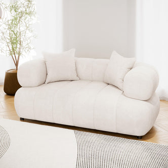 Samira 2 Seater Velvet Lounge in Cream, featuring softly rounded upholstery, cylindrical bolster arms, and two matching throw pillows, sits elegantly on a subtle patterned rug, exuding a modern, minimalistic vibe.
