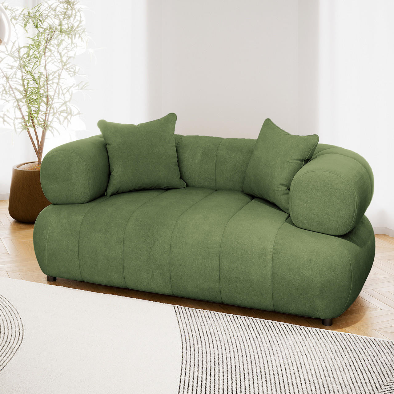 Samira 2 Seater Velvet Lounge in Prasinous Green, featuring a low-slung design, chunky rounded arms, and two matching pillows, set against sheer curtains and a leafy plant on parquet flooring.
