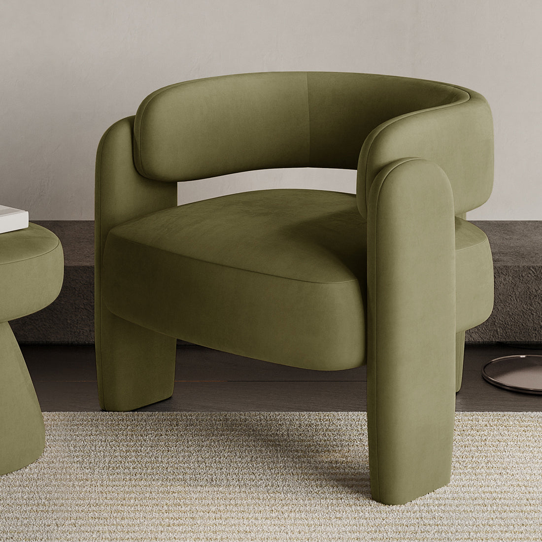 Seto Velvet Curved Occasional Accent Chair – Sage features an olive-green fabric, curved backrest, rounded arm supports, and thick legs on a woven rug, with a matching side table nearby.