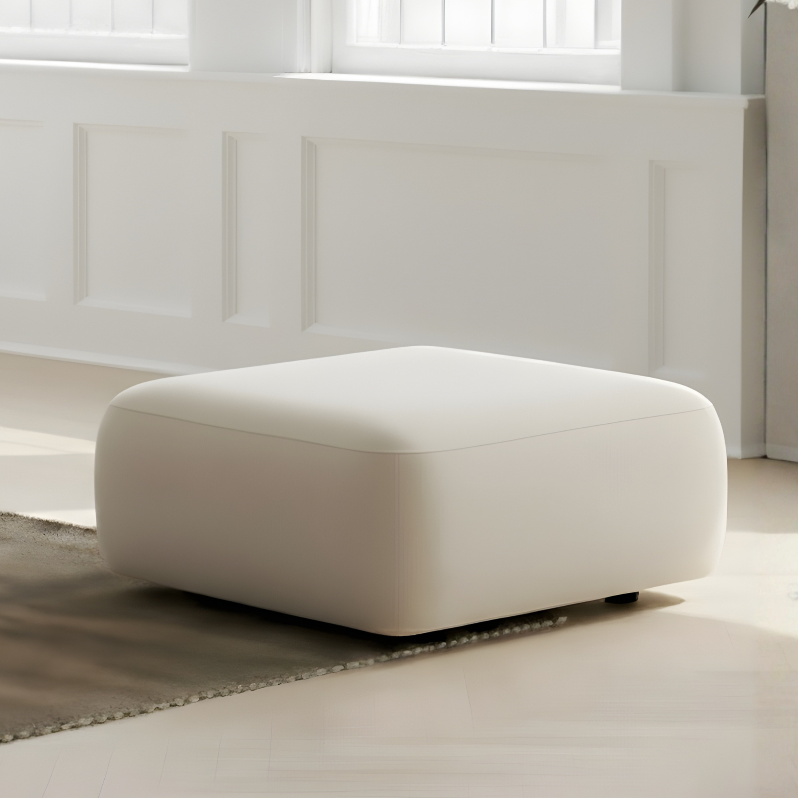 Soraya Velvet Fabric Ottoman in Cream White, featuring a square shape and light cream upholstery, placed on a neutral rug in a sunlit corner with white paneled walls and windows.