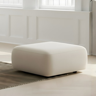 Soraya Velvet Fabric Ottoman in Cream White, featuring a square shape and light cream upholstery, placed on a neutral rug in a sunlit corner with white paneled walls and windows.