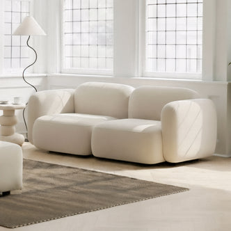 Soraya 2 Seater Velvet Sectional Sofa in Cream White, featuring a low-slung, cloud-like design with softly rounded edges, highlighted in a bright, minimalist living area with large windows and a sculptural side table.