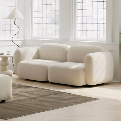 Soraya 2 Seater Velvet Sectional Sofa in Cream White, featuring a low-slung, cloud-like design with softly rounded edges, highlighted in a bright, minimalist living area with large windows and a sculptural side table.