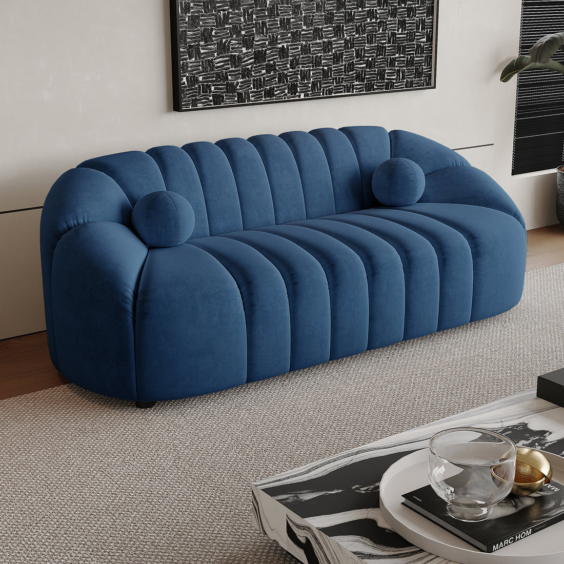 Trina 3 Seater Tufted Velvet Sofa in Blue features plush channel tufting, gently rounded arms, and two bolster pillows, set in a modern living room with a marble-pattern coffee table and abstract wall art.