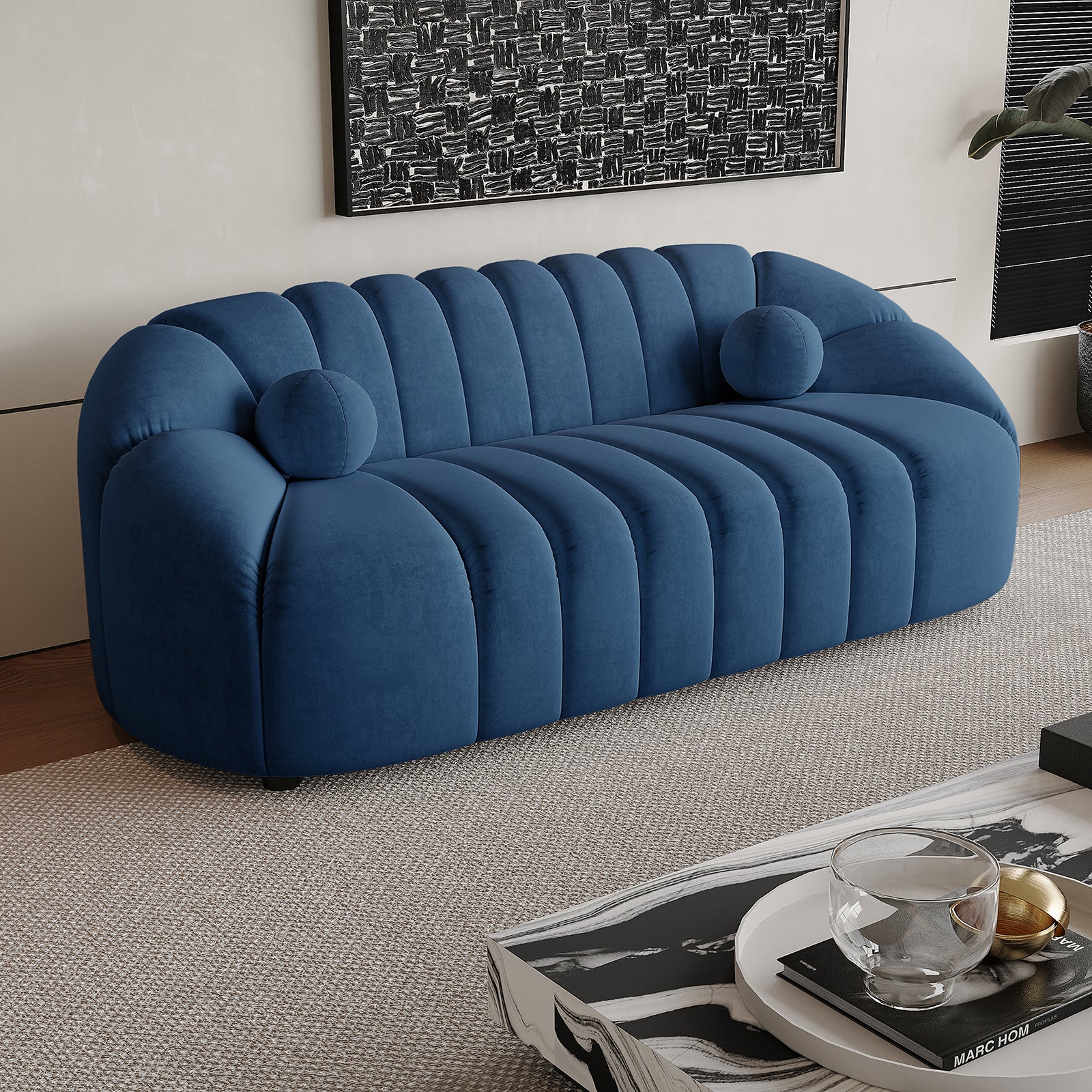 Trina 3 Seater Tufted Velvet Sofa in Blue features plush channel tufting, gently rounded arms, and two bolster pillows, set in a modern living room with a marble-pattern coffee table and abstract wall art.