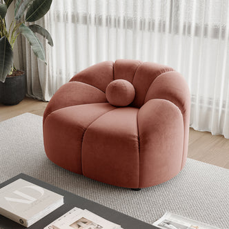 Trina Velvet Sofa Chair with Ball Pillow in Blush features petal-like segments and a round bolster cushion, set on a light-gray woven rug, complemented by a nearby dark coffee table and tall plant.