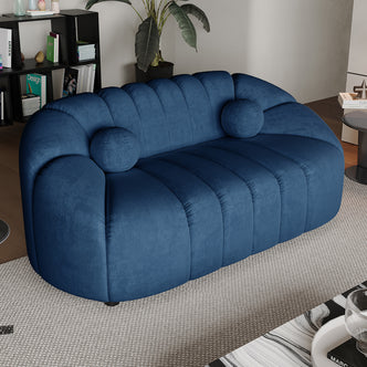 Trina 2 Seater Tufted Curved Velvet Sofa in Blue features plush upholstery, channel tufting, matching bolster pillows on a light-beige rug, complemented by a black shelving unit and a side table.