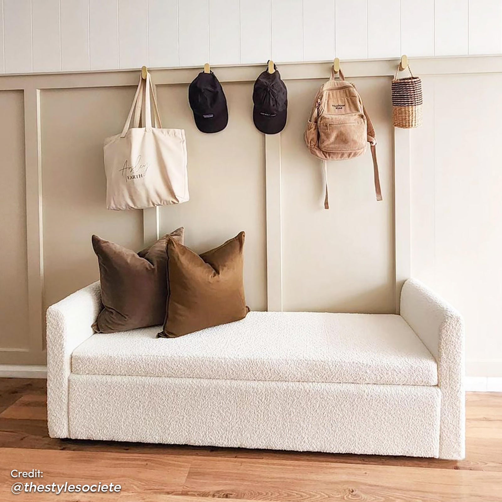 Zamira Large Boucle Storage Ottoman Bench in Cream White, featuring a creamy bouclé fabric with rust-brown pillows, set against a paneled wall with wall hooks holding various items.