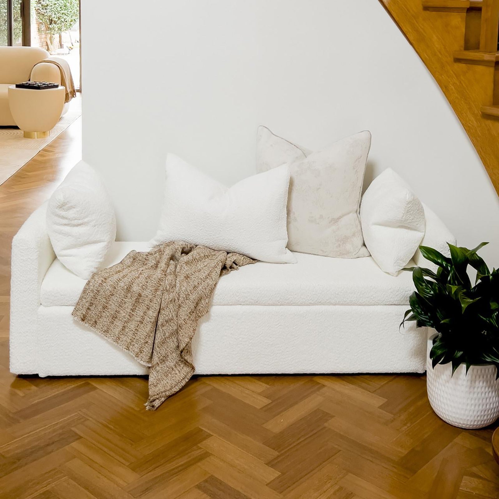 Zamira Large Boucle Storage Ottoman Bench in Cream White under staircase, styled with white throw pillows and taupe knit throw. Featured against warm herringbone floor and adjacent to a green houseplant.