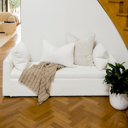 Zamira Large Boucle Storage Ottoman Bench in Cream White under staircase, styled with white throw pillows and taupe knit throw. Featured against warm herringbone floor and adjacent to a green houseplant.