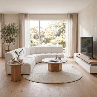 Contemporary Airbnb living room featuring a curved white bouclé modular sectional sofa, an organic-shaped cream rug, a round marble-topped coffee table with a wooden base, a matching side table, and a wall-mounted TV over a low white and wood media console.
