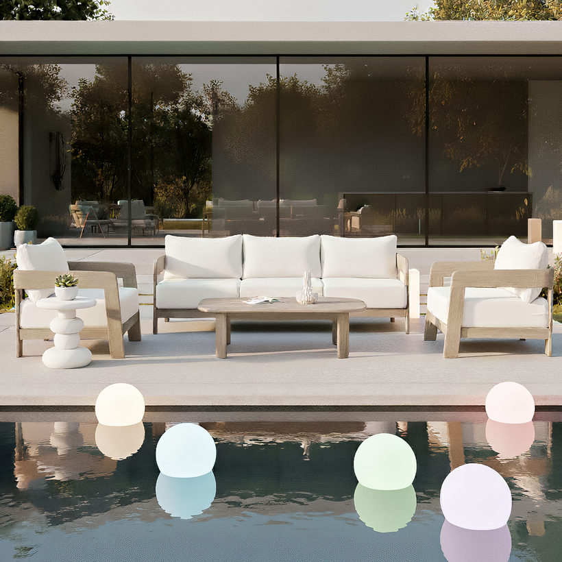 Modern outdoor lounge setting with a light wood sofa, armchairs and coffee table on a poolside terrace, styled with white cushions and glowing floating orb lights in the water, in front of a contemporary glass-walled home.