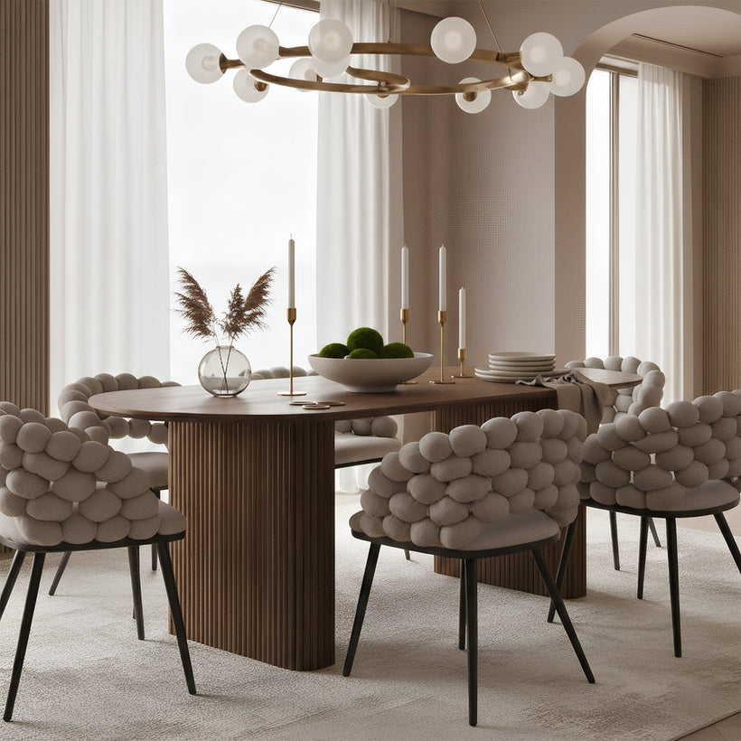A modern dining room with a warm, neutral palette. A rounded wooden dining table with a ribbed pedestal base is surrounded by upholstered chairs featuring chunky, bubble-like cushions and black tapered legs. On the table are slim gold candlesticks with white candles, a shallow bowl filled with green moss spheres, stacked plates, and a clear glass vase holding dried grasses. Soft daylight filters through sheer curtains, creating a calm, minimalist atmosphere.
