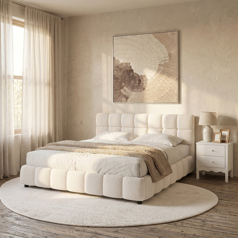 Cream bouclé upholstered bed frame with a padded square-panel headboard and matching cushioned base, styled in a warm neutral bedroom with a round cream rug, white bedside table, soft linen curtains, and textured wall art illuminated by natural sunlight.