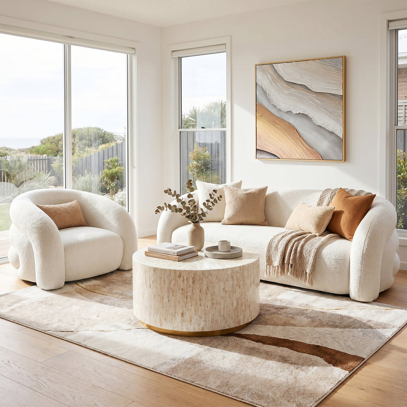 Contemporary living room featuring a curved white bouclé sofa and matching accent chair, styled with neutral-toned cushions around a round marble-look coffee table. The space is complemented by a textured abstract rug, warm wood flooring, large sunlit windows, and modern abstract wall art in soft beige and grey tones.