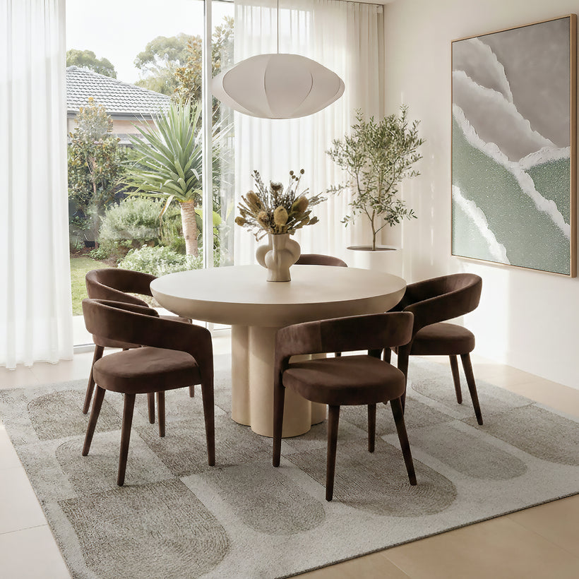 Modern dining room featuring a round light-toned dining table with six brown upholstered chairs, a sculptural pendant light overhead, a neutral patterned rug, soft sheer curtains, indoor plants, and abstract wall art, with natural light coming through large glass doors.