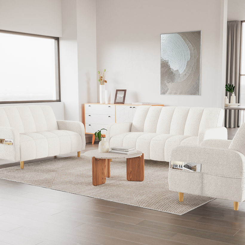 Bright, modern living room with matching cream upholstered sofas featuring channel tufting, arranged around a small round wooden coffee table on a textured neutral rug. The space includes minimalist wall art, a light wood sideboard with decor, large windows, and soft natural light for a clean, cozy aesthetic.