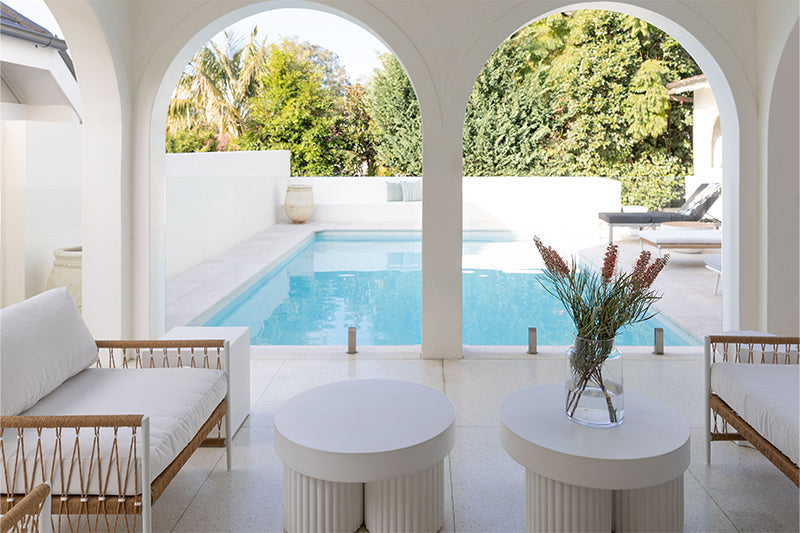 Luxo Living outdoor sofa and concrete coffee tables styled in beautiful backyard setting with pool and arch detail architecture with sunbeds in the background