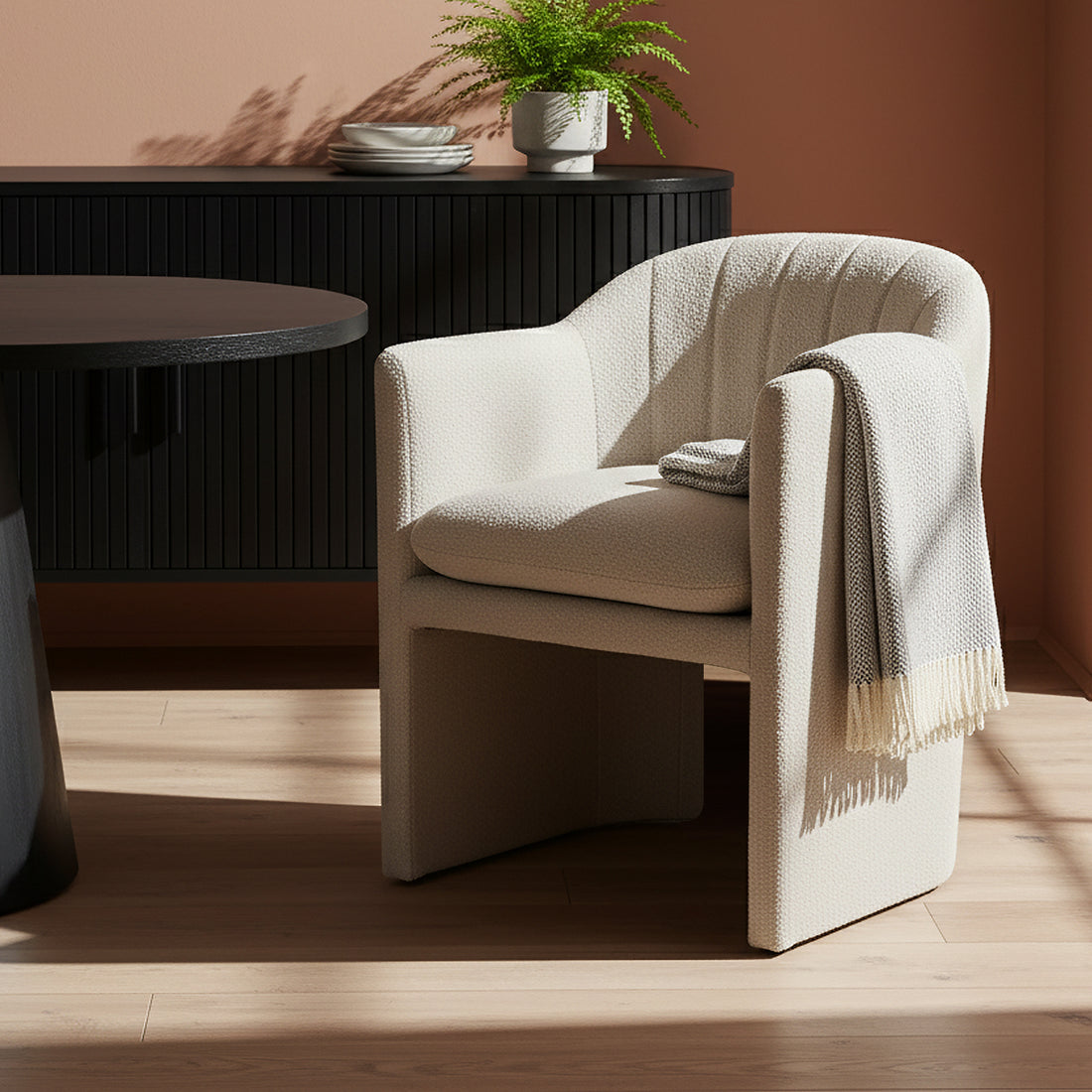 Liora Bouclé Curved Accent Armchair in Beige, featuring a pale textured fabric, is nestled in a softly lit corner with a fringed throw and a nearby black side table.