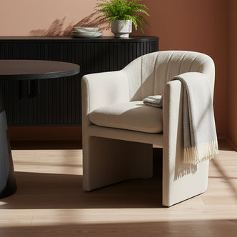 Liora Bouclé Curved Accent Armchair in Beige, featuring a pale textured fabric, is nestled in a softly lit corner with a fringed throw and a nearby black side table.