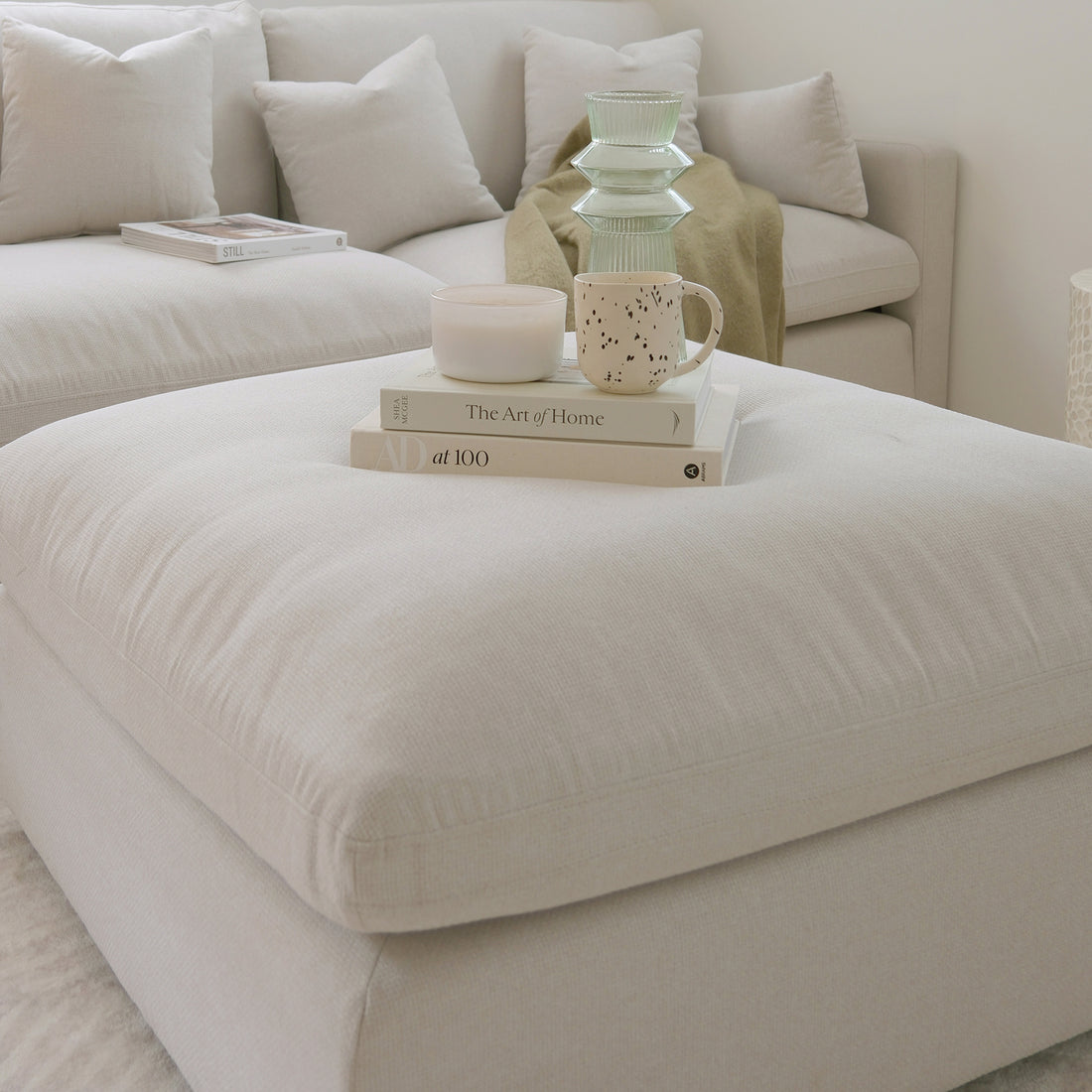 Partial view of a light-colored sectional sofa and ottoman styled with books, a candle, and a mug in a living room.