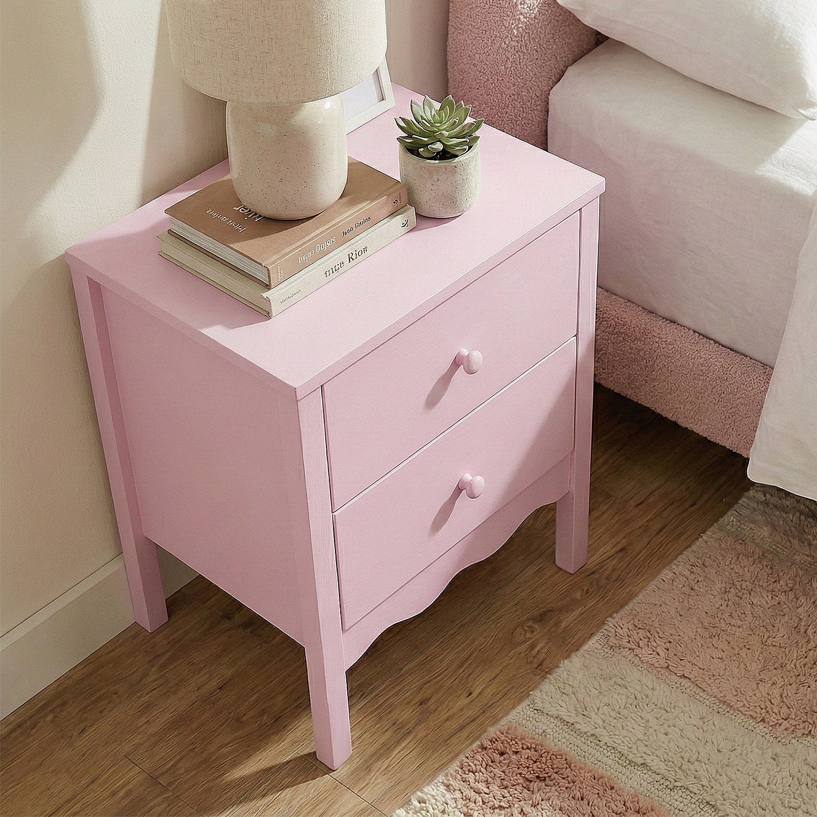 Pink two-drawer nightstand in a bright bedroom setting with lamp, books, and potted plant