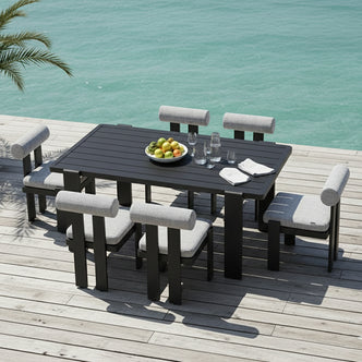 Positano 6 Seater Outdoor Dining Set in Black & Grey on a deck by turquoise water, featuring black frames, grey cushions, and a table with fruit, glasses, and napkins, hinting at a seaside setting.