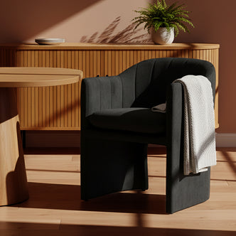 Liora Velvet Curved Accent Armchair in Black features sculptural lines and plush upholstery, with a light throw draped over it, set against a warm wood floor and sideboard.