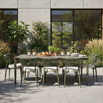 Cabria 9 Piece Sintered Stone Outdoor Dining Set features a rectangular table and eight woven rope chairs with cushions, set with fruits, herbs, and candles on a patio amidst lush landscaping.