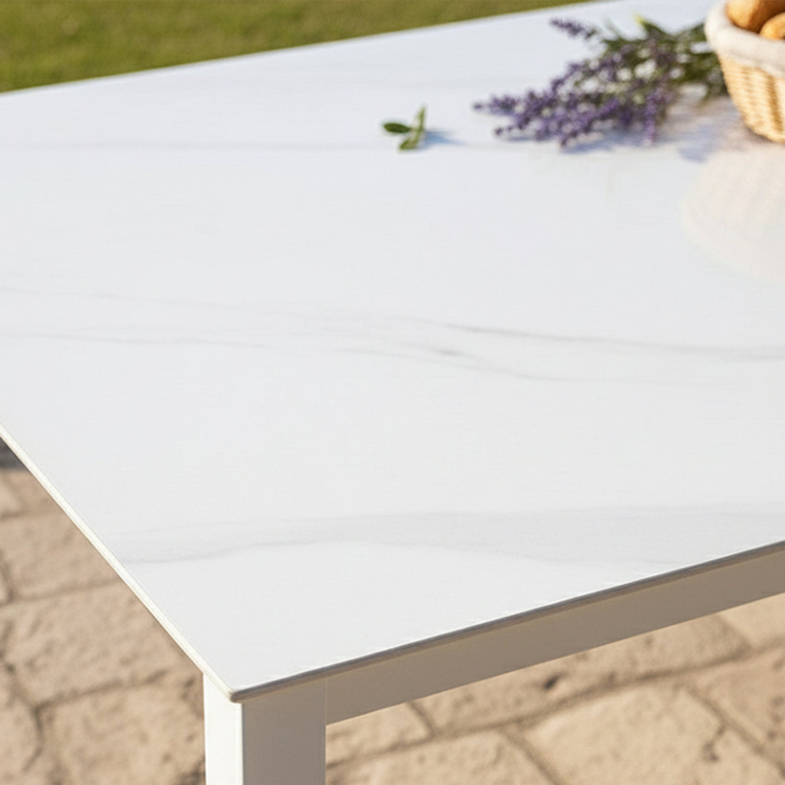 Cabria 9 Piece Sintered Stone Outdoor Dining Set showcased with a white marble-style tabletop on a patio, complemented by a wicker basket with rolls and a lavender sprig, highlighting elegance and durability.